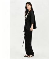 Full body shot of a women's black silk tunic top with a wrap front and tied waist, showcasing the relaxed fit and elegant drape of the 100% silk fabric, styled with black wide-leg pants and sandals.