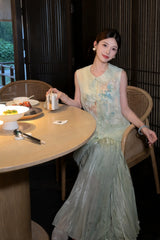 Sitting model in a green silk-blend vest with floral embroidery and pearl buttons, holding a small object, showcasing the relaxed fit and elegant drape.