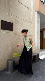 Side view of a woman in a jade floral brocade Cheongsam vest, looking down while holding a bright green bag, highlighting the sleeveless design and silky drape.