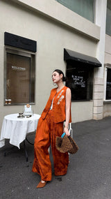 Model standing by stone wall in a rust orange Cheongsam set, looking to the side, with white floral embroidery on the vest.